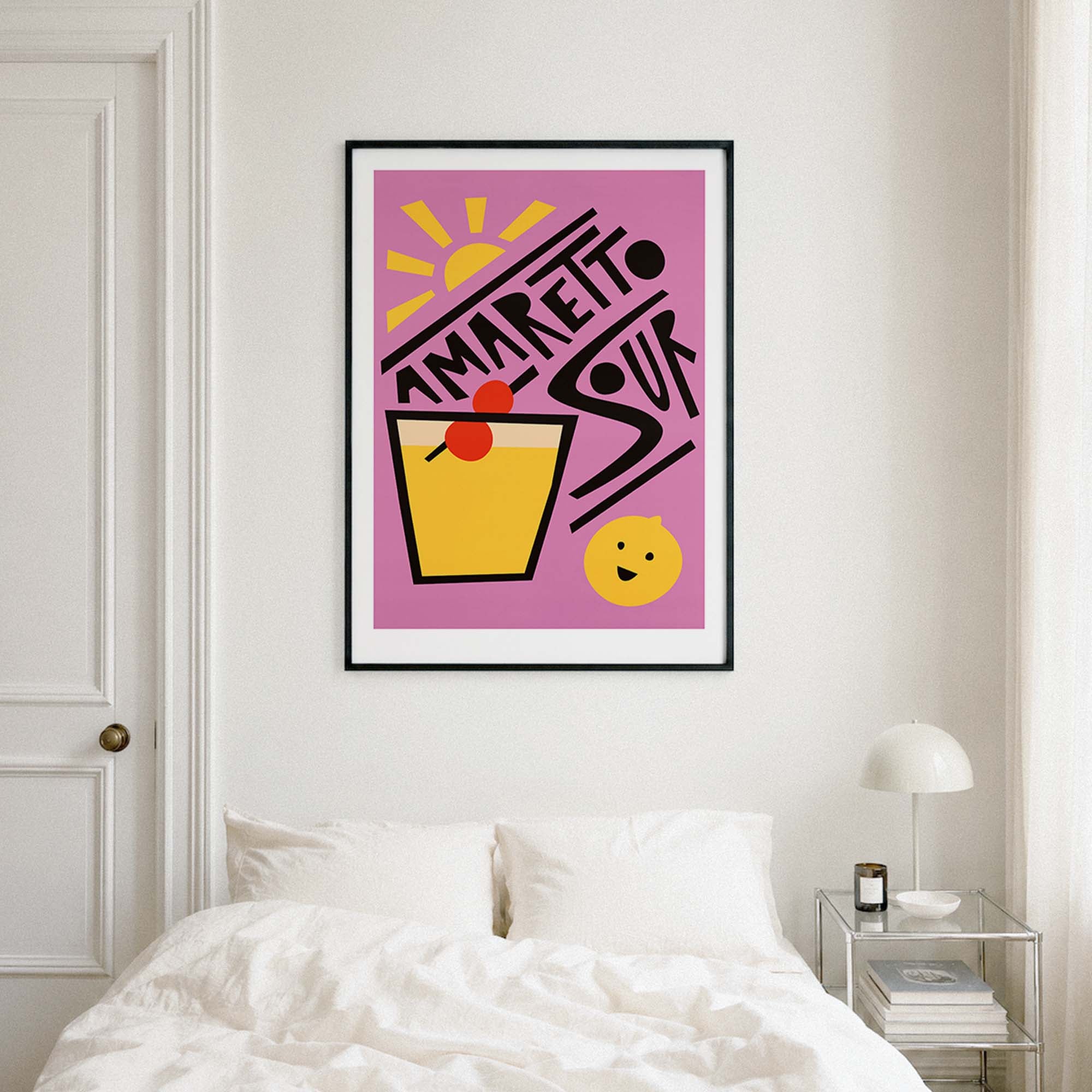 A framed art print featuring a retro-style 'Amaretto Sour' cocktail illustration in pink, yellow, black, and red, displayed on a bedroom wall above a bed.