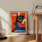 Aquarius print framed on the floor next to a pale wood desk and books in a clean, modern nook.