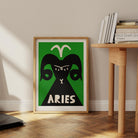 Aries print framed on the floor beside a minimalist wood desk with stacked books in a neutral-toned interior.