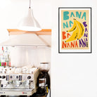 Illustrated banana poster adding pop-art flair to a kitchen corner beside a coffee station.

