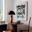 Brighton cityscape print hung above a desk nook with a black dome lamp, blending mid-century style with coastal flair in a relaxed home office setting.
