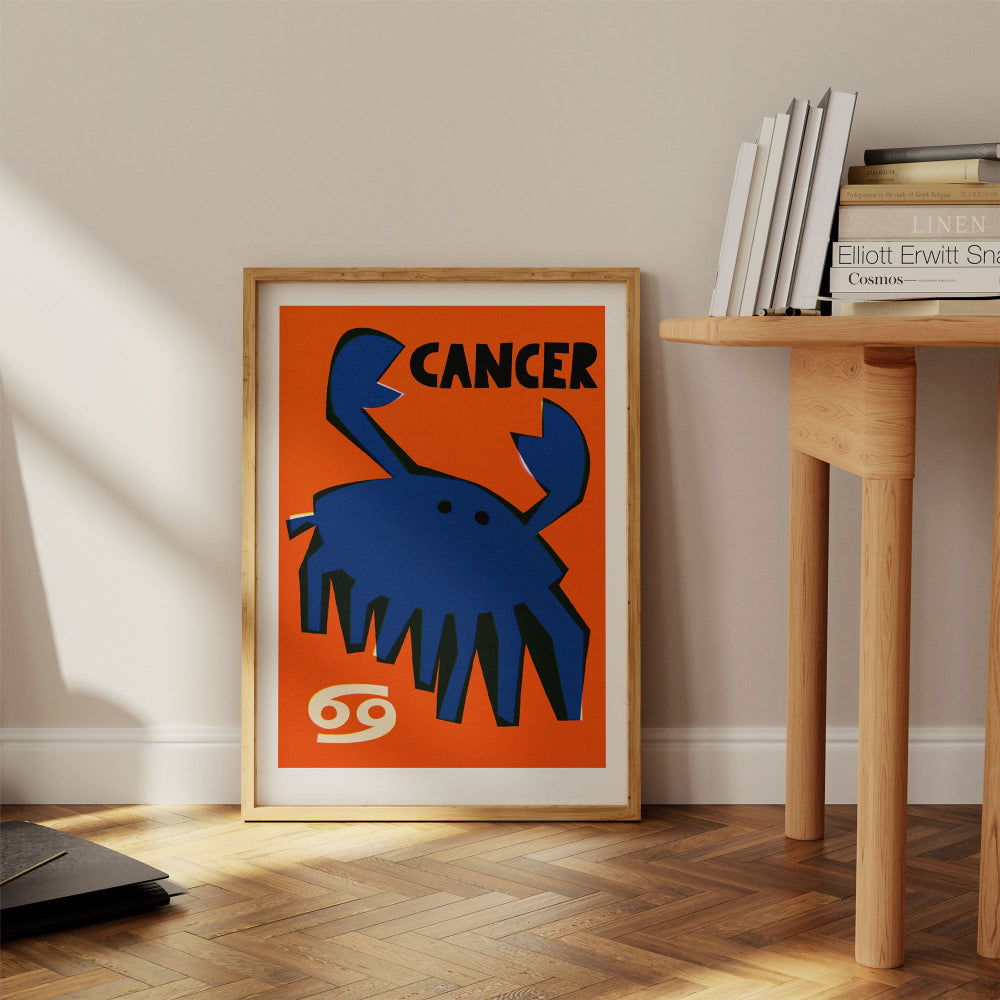 Cancer print framed on the floor next to a pale wood desk with stacked books in a quiet reading space.