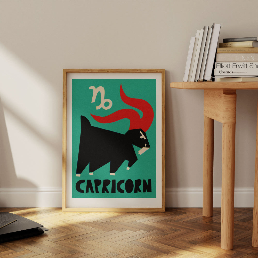 Capricorn print framed on the floor beside a pale wood desk with stacked books in a minimal home space.