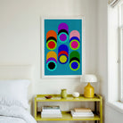 A framed abstract art print featuring colorful geometric circles on a teal background, hanging on a white wall above a yellow shelf in a bright bedroom setting.