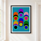 Framed abstract geometric wall art hanging on a white wall next to a window. The print features bold, colorful cylinder shapes in multi colours against. light blue background
