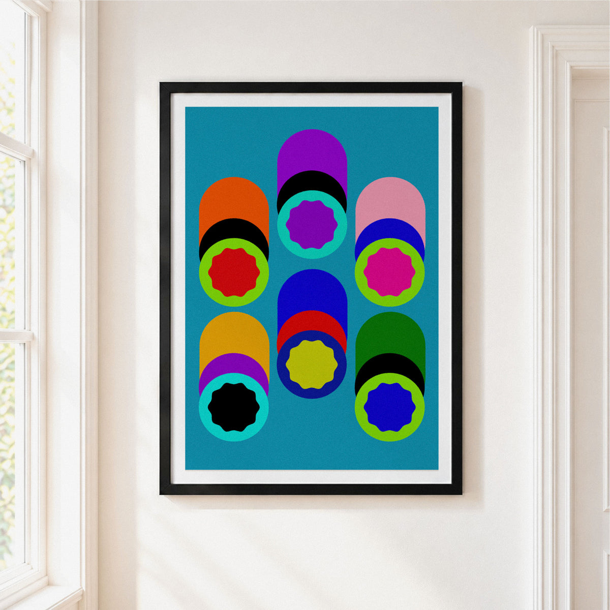 Framed abstract geometric wall art hanging on a white wall next to a window. The print features bold, colorful cylinder shapes in multi colours against. light blue background
