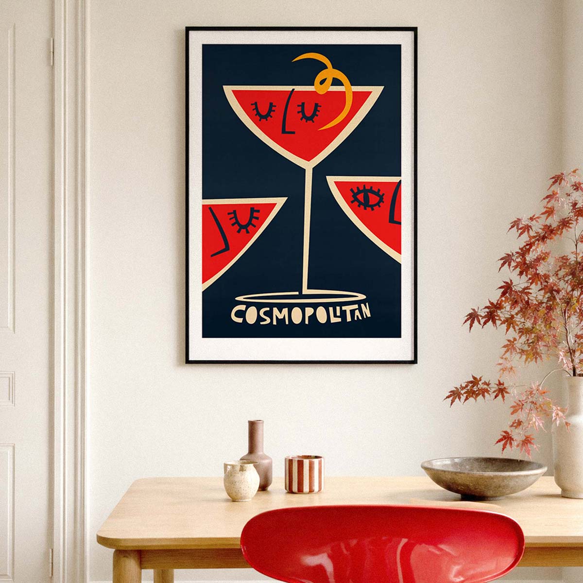 A framed art print featuring a stylized cocktail illustration with the word 'COSMOPOLITAN' at the bottom, displayed on a light beige wall above a wooden table with decorative vases and a red chair.