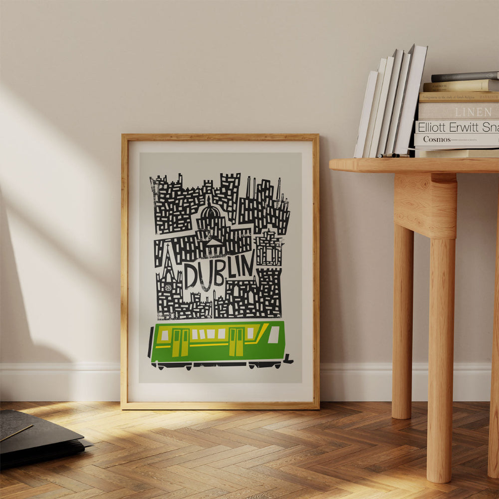 Framed Dublin city print propped against a wall on a wooden floor, showcasing the charming chaos of Dublin’s skyline and the vibrant green LUAS tram below.