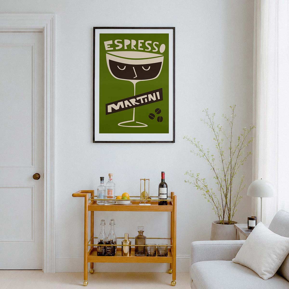 A framed vintage-style wall art print featuring an espresso martini glass design with the words 'ESPRESSO MARTINI' in a green, cream, and dark brown palette, hanging on a white wall above a wooden bar cart in a styled living room.
