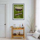 A framed vintage-style wall art print featuring an espresso martini glass design with the words 'ESPRESSO MARTINI' in a green, cream, and dark brown palette, hanging on a white wall above a wooden bar cart in a styled living room.