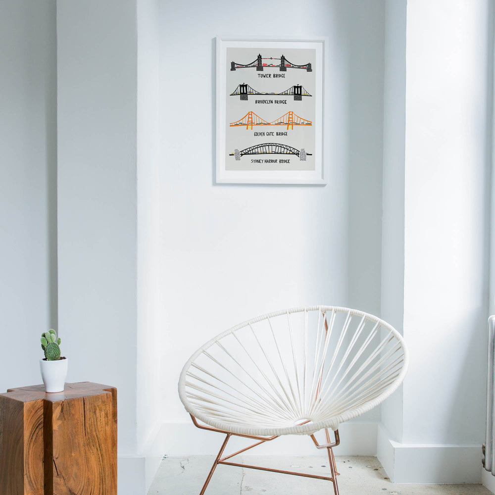 Framed famous bridges print hanging in a minimal white space with natural light and a rattan chair, giving a travel-inspired, editorial feel.