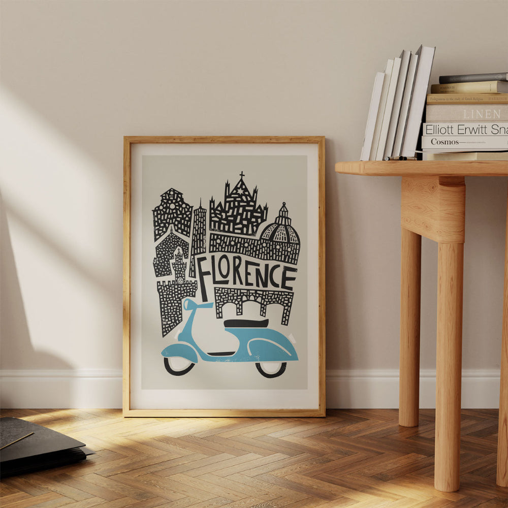 Florence cityscape print leaning casually against a wall, with bold black landmarks rising above a vintage blue motorbike in a modern wooden-floored setting.