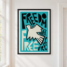 A framed art print featuring a stylized white dove and the lettering typogrpahy 'FREEDOM' on a turquoise background, hanging on a white wall next to a window.