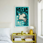 A framed art print featuring a stylized white dove and the lettering typography 'FREEDOM' on a teal background, hanging on a white wall above a yellow bedside table in a bright bedroom.