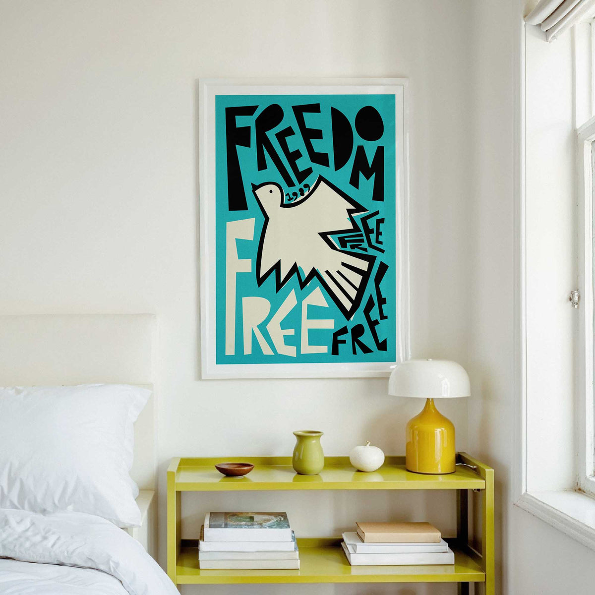 A framed art print featuring a stylized white dove and the lettering typography 'FREEDOM' on a teal background, hanging on a white wall above a yellow bedside table in a bright bedroom.