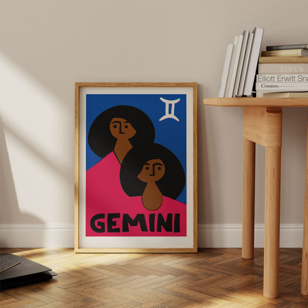 Gemini print framed on the floor beside a pale wood desk and books in a minimalist interior.
