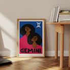 Gemini print framed on the floor beside a pale wood desk and books in a minimalist interior.