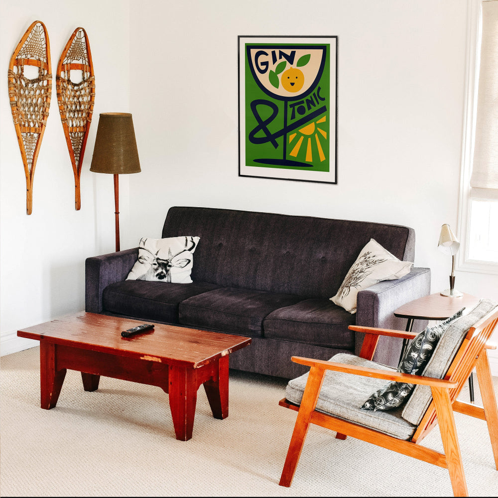 Gin & Tonic poster framed above a dark sofa in a cosy mid-century living space with vintage skis and warm wood accents.