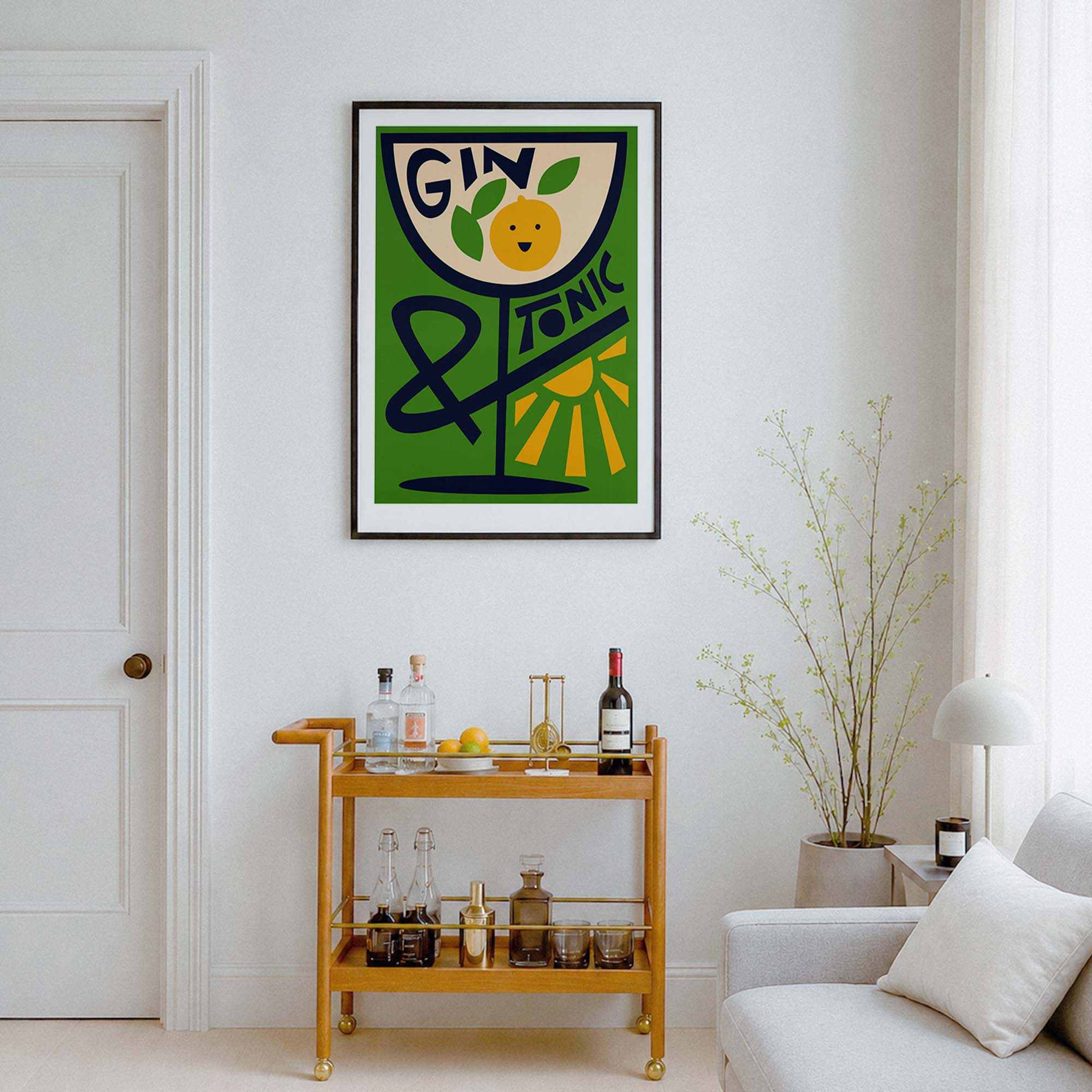 A framed vertical art print featuring a stylized green and cream cocktail glass with the words 'GIN & TONIC' and a lemon illustration, displayed on a white wall above a wooden bar cart in a modern living room setting.