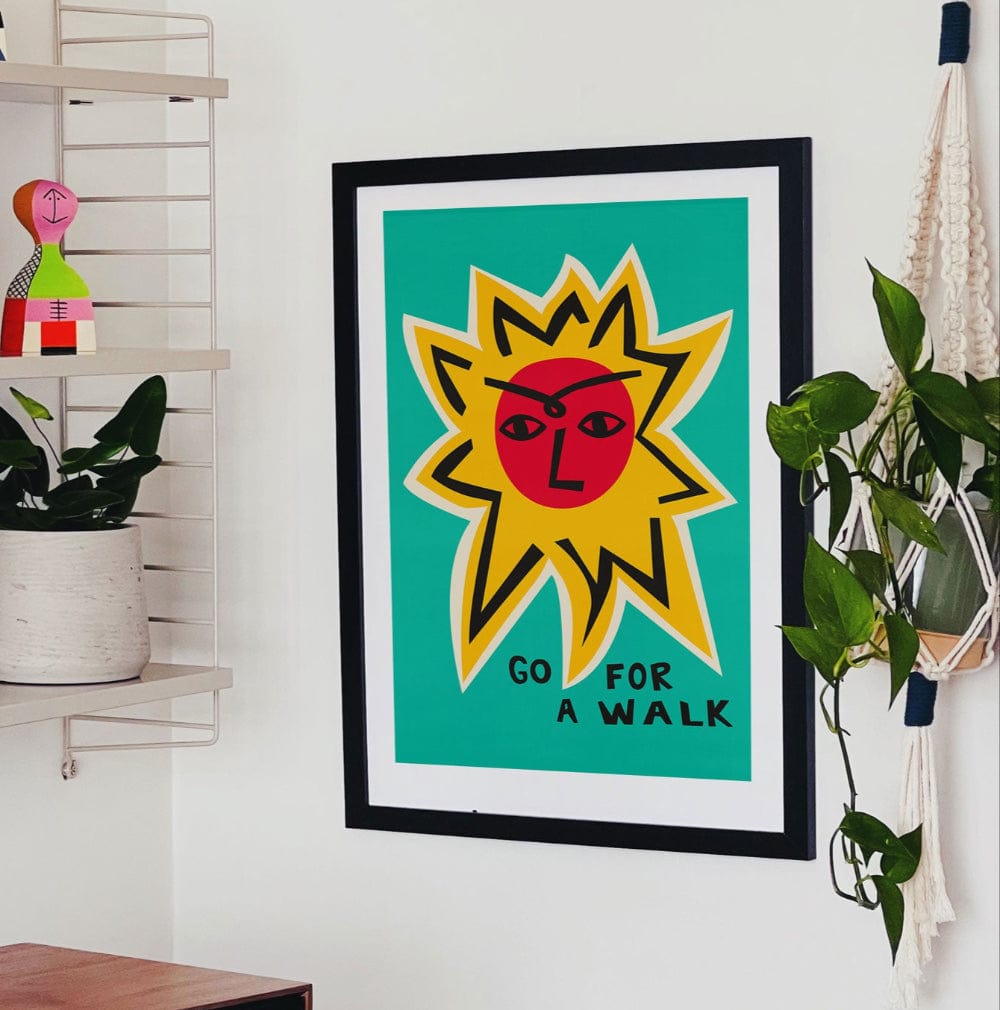 Go For a Walk poster framed above a desk in a colourful studio corner with houseplants and shelving.