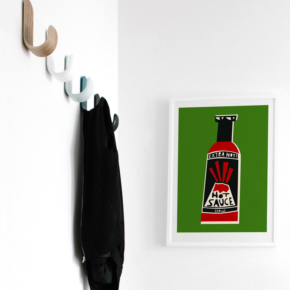 Hot sauce artwork framed on a white wall beside coat hooks and a hanging black jacket.