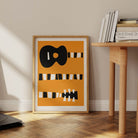 Framed guitar illustration leaning against a light wall beside a wooden desk and stack of books