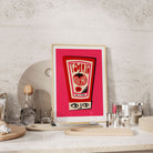 Ketchup bottle print framed on a kitchen shelf with stone backdrop and scattered barware.
