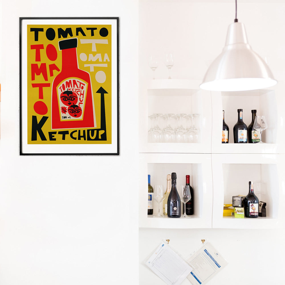 Framed Tomato Ketchup print placed on a crisp white wall beside open kitchen shelves, its mustard yellow background making the red bottle pop with bold contrast.
