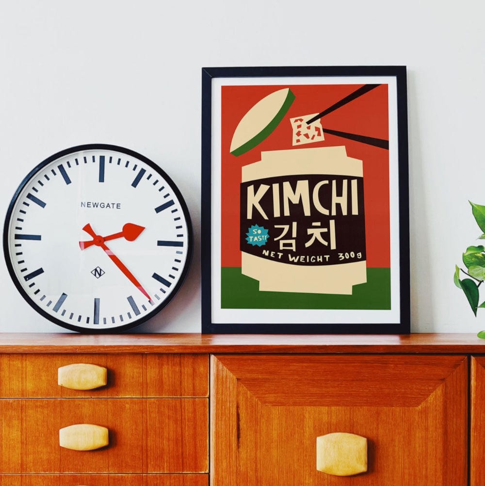 Kimchi poster framed on a wooden sideboard beside a large clock and indoor plant in a retro-modern room.