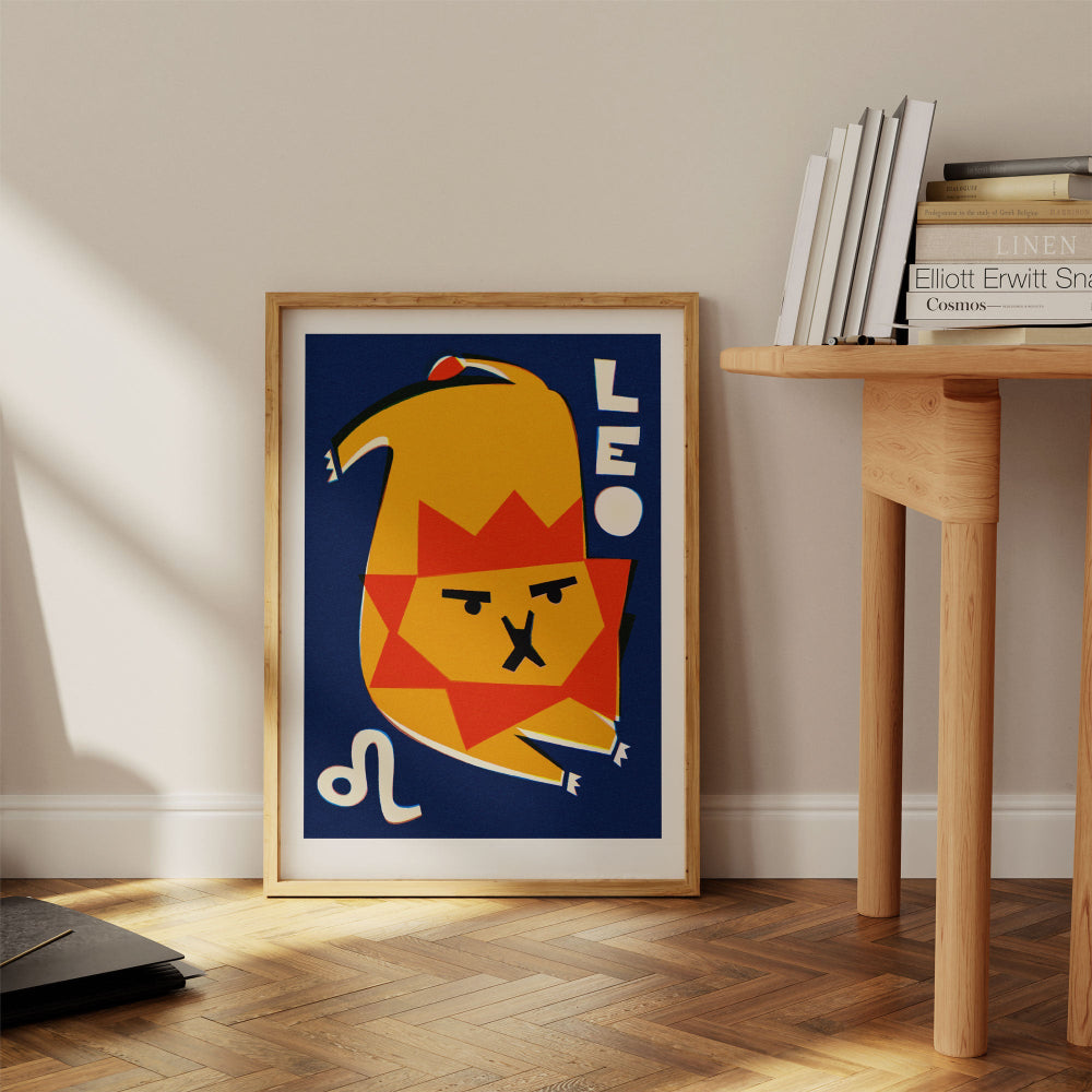 Leo zodiac print framed on the floor next to a pale wooden desk with stacked books in a warm-toned interior.
