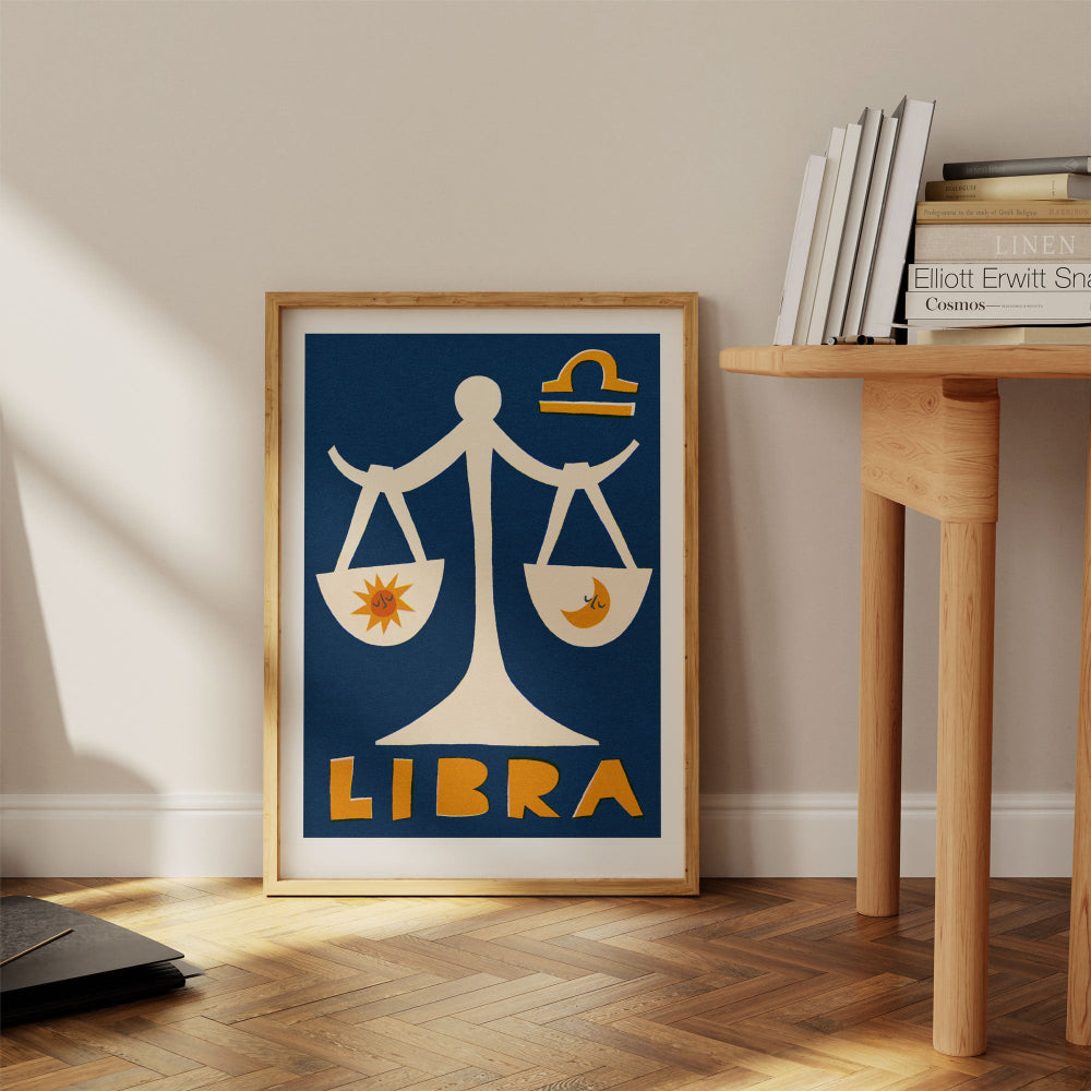 Libra print framed on the floor next to a wooden desk with stacked books and pale wall.