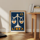 Libra print framed on the floor next to a wooden desk with stacked books and pale wall.