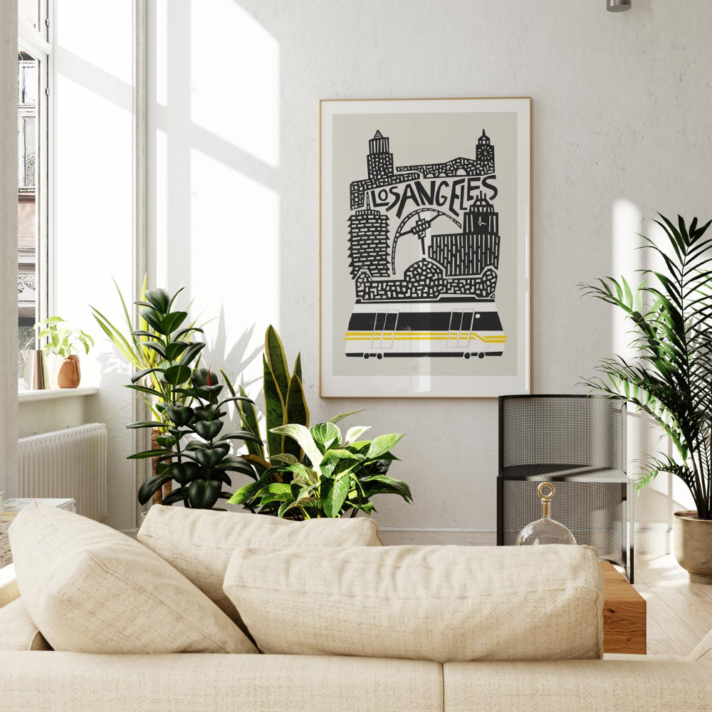 Framed LA art print hanging in a bright living room, blending modern cityscape artwork with calm, neutral home decor and leafy indoor plants.