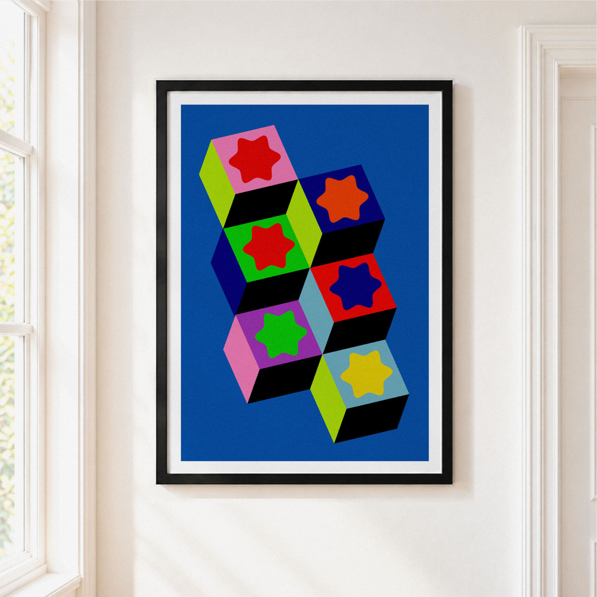 Framed abstract geometric wall art hanging on a white wall next to a window. The print features bold, colorful 3D block shapes in red, lime green, black, and navy blue on a lavender background, enclosed in a black frame with a white mat.
