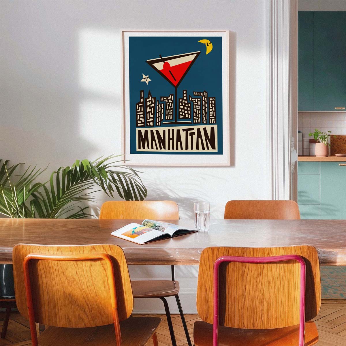 A framed art print featuring a retro-style cocktail and the word 'MANHATTAN' hangs on a white wall above a wooden dining table with mid-century modern chairs in a bright, modern kitchen setting.