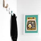 Mayonnaise print framed in a bright, minimalist space with wall hooks and a black coat.