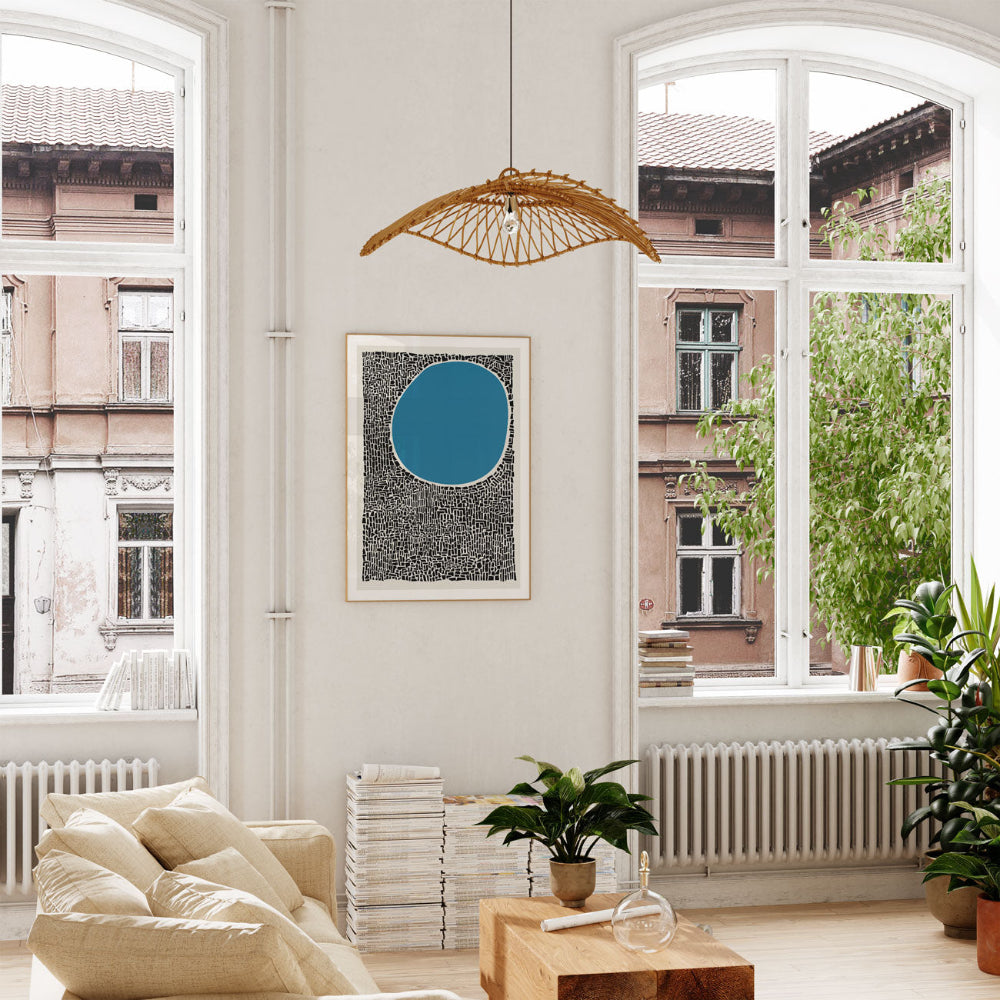 Framed abstract blue circle print hung near tall arched windows, surrounded by greenery, basking in natural light in a neutral, airy interior.