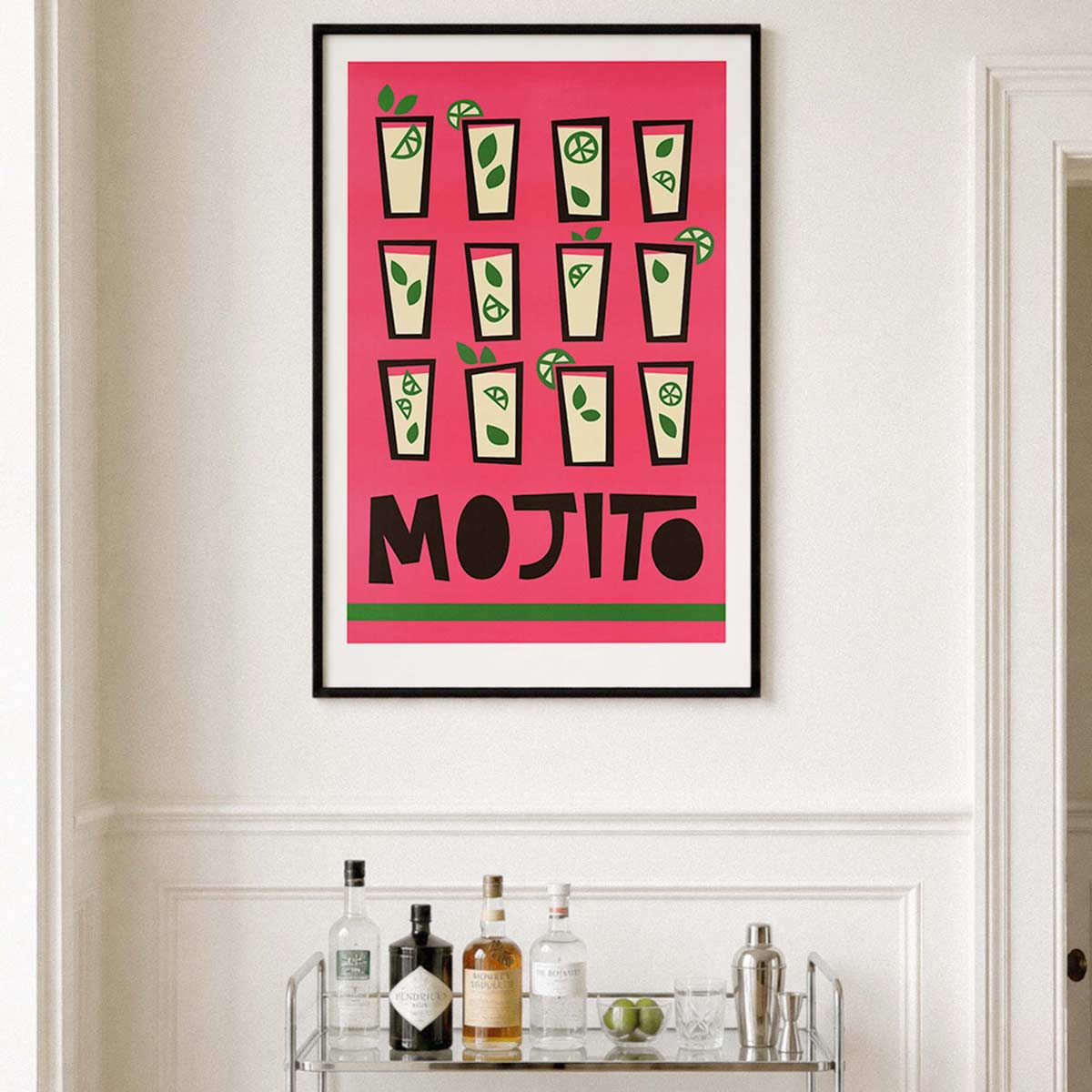 A framed art print featuring a retro Mojito cocktail design with illustrations of mojitos and the word 'MOJITO' in bold black letters on a bright pink background, hanging on a white wall above a bar cart with bottles.