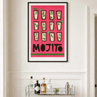 A framed art print featuring a retro Mojito cocktail design with illustrations of mojitos and the word 'MOJITO' in bold black letters on a bright pink background, hanging on a white wall above a bar cart with bottles.