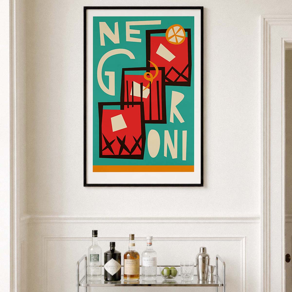 A framed art print featuring a retro-style illustration of three red and black cocktails with cream lettering on a teal background, displayed on a white wall above a bar cart with bottles.