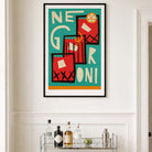 A framed art print featuring a retro-style illustration of three red and black cocktails with cream lettering on a teal background, displayed on a white wall above a bar cart with bottles.