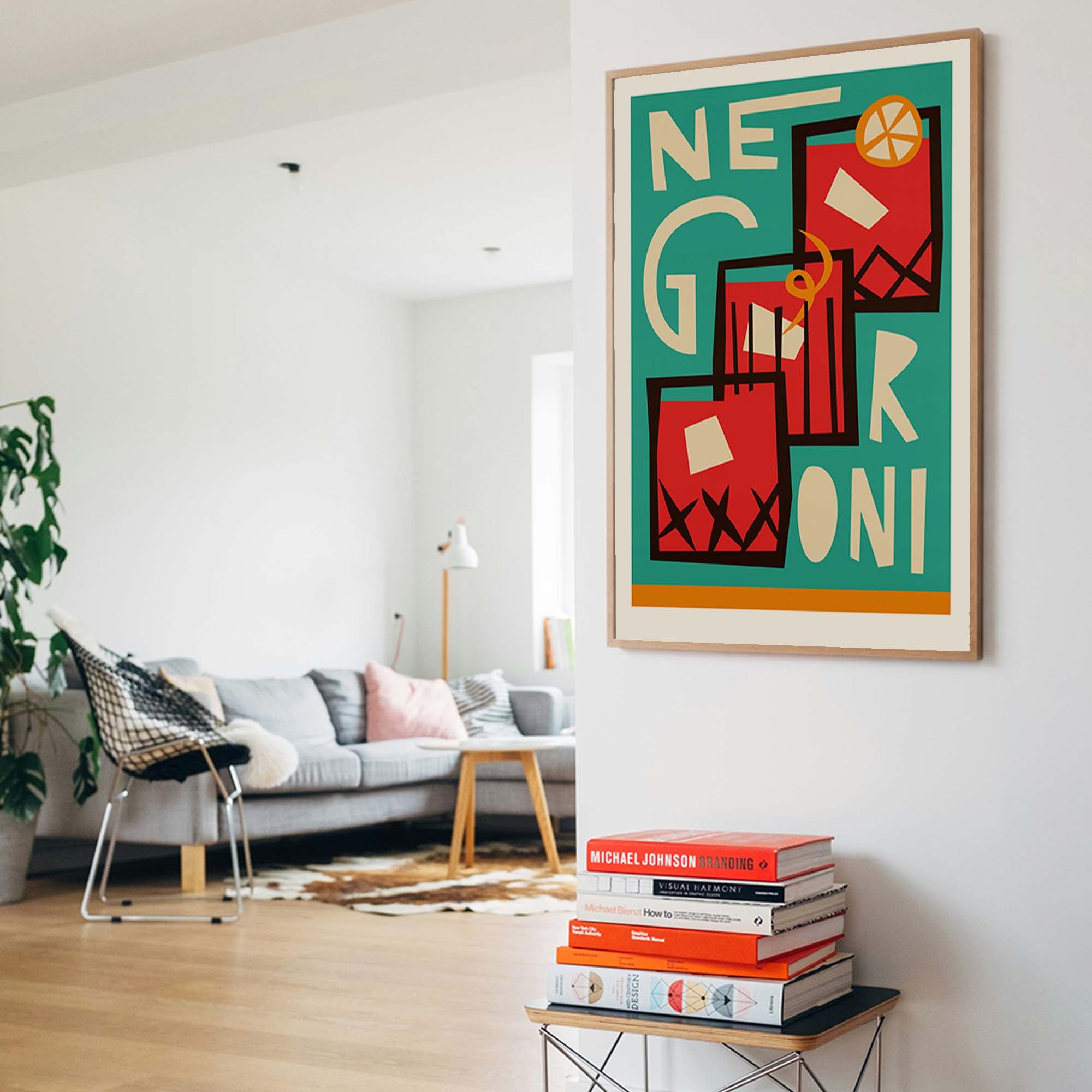 A framed art print featuring a retro-style illustration of Neer Gini cocktails hangs on a white wall in a modern, sunlit living room. The print has a teal background with red, orange, and cream geometric drink illustrations and bold cream lettering.