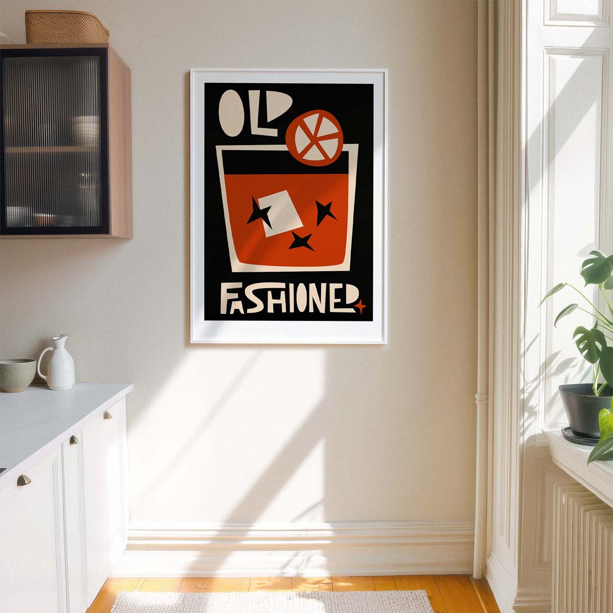 A framed vintage-style cocktail poster hanging on a beige wall in a bright kitchen or dining area. The poster features bold retro typography reading 'OLD FASHIONED' with an orange drink glass, white garnish, and black background.