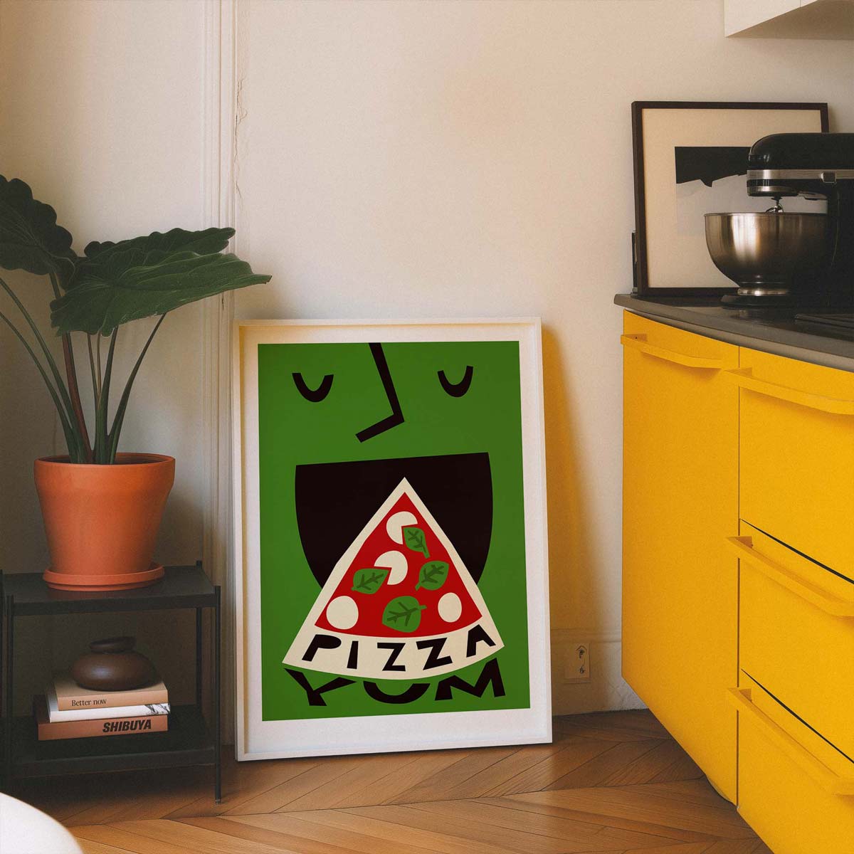 A framed art print featuring a playful cartoon face with a pizza slice for a mouth and the word 'PIZZA YUM' below, leaning against a wall in a kitchen setting next to a yellow cabinet and a potted plant.