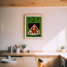 A framed art print featuring a stylized green face eating a slice of pizza with the word 'PIZZA' inside, hanging on a light beige wall above a wooden kitchen counter.