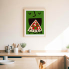 A framed art print featuring a stylized green face eating a slice of pizza with the word 'PIZZA' inside, hanging on a light beige wall above a wooden kitchen counter.