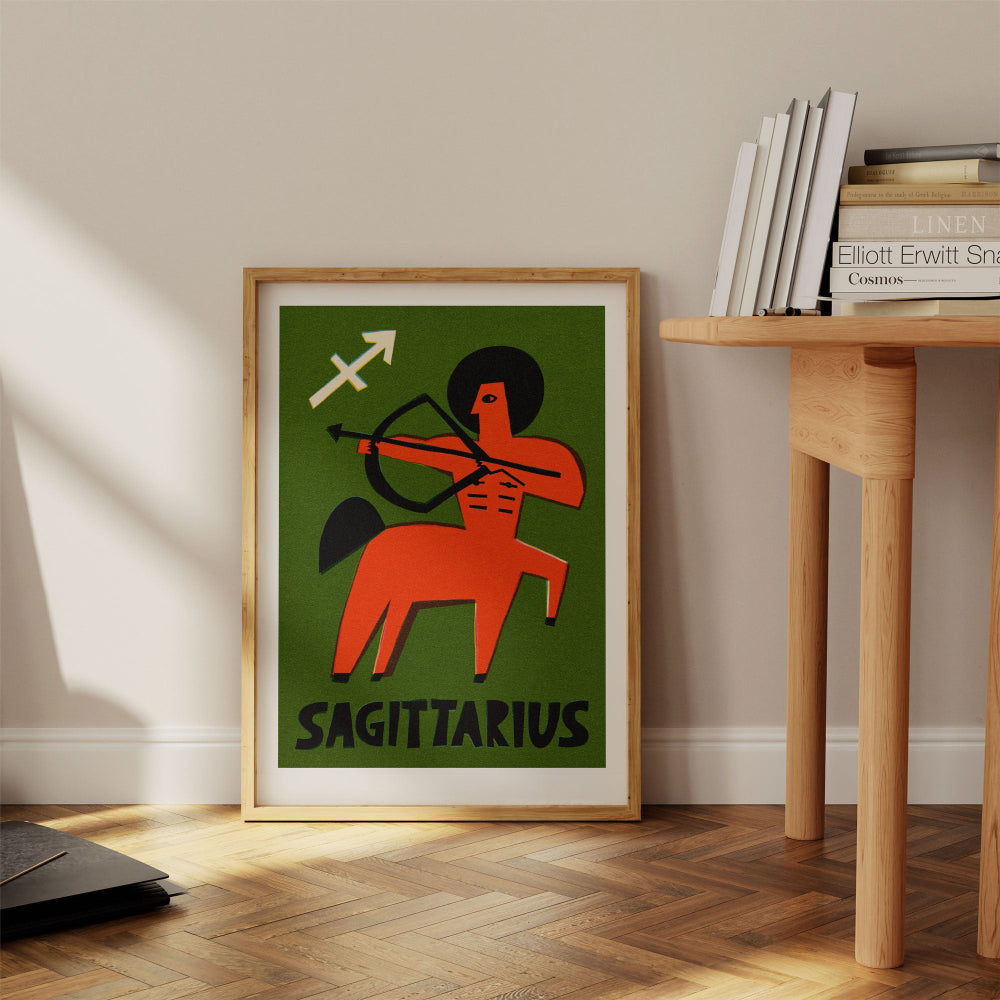 Sagittarius print framed on the floor beside a pale wood desk with stacked books and warm light.
