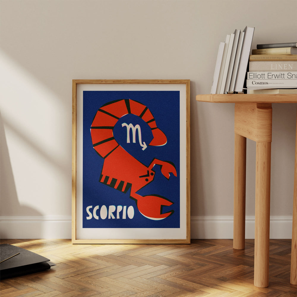 Scorpio zodiac print framed on the floor next to a wood desk and books in a warm, casual interior.