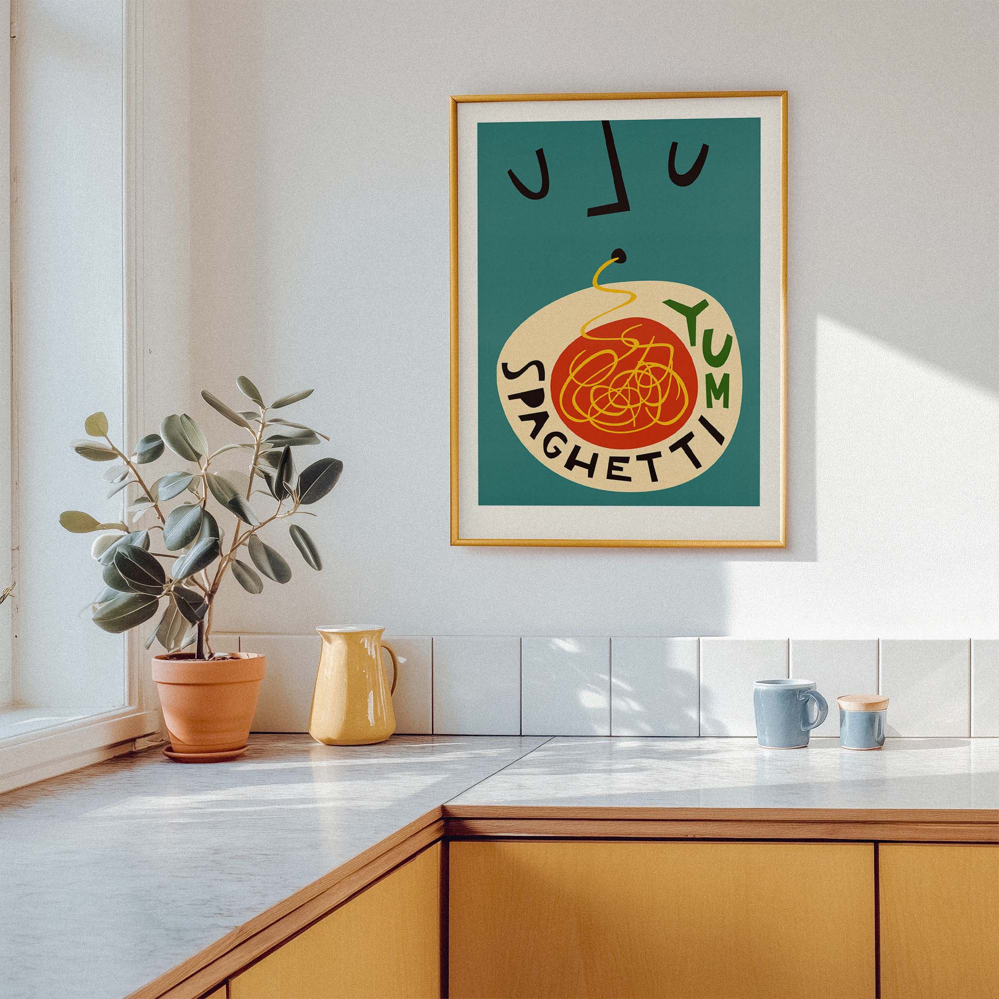 A framed art print featuring a retro-style illustration of spaghetti with the words 'JUM SPAGHETTI' on a teal background, hanging on a white kitchen wall above a counter with a potted plant and mugs.