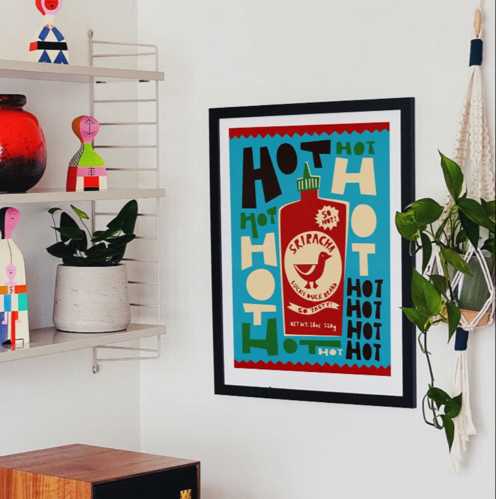 Sriracha Lucky Duck artwork framed on a white wall beside open shelving, plants, and colourful decor.